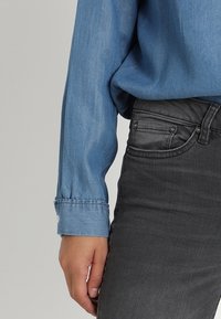 Camisa de mezclilla azul de manga larga con puños abotonados, combinada con jeans gris oscuro con bolsillos frontales y tachuelas metálicas. Tejido texturizado.
