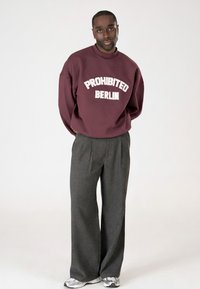 Muž stojí s rukama za zády, oblečený v vínovém mikině s nápisem "PROHIBITED BERLIN", šedých širokých kalhotách a bílých teniskách na čistém pozadí.