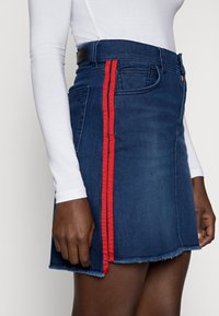 Džínová sukně v tmavě modré barvě se třepícími okraji, se červenými bočními pruhy a předním zipem. Obsahuje přední kapsy a pásek s logem.