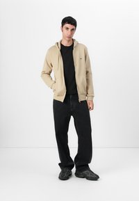 Felpa beige con zip in morbido tessuto, t-shirt nera sotto, abbinata a jeans neri larghi e sneakers nere con texture.