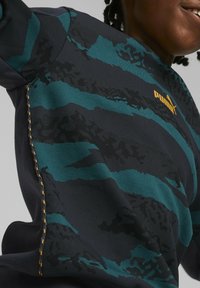 Felpa blu scuro con motivi a strisce turchesi e dettagli di camuffamento sottili. Logo dorato "PUMA" e ricamo testurizzato in evidenza.