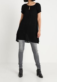 Tunique noire avec un col en trou de serrure, manches courtes bouffantes et plis, associée à un jean gris ajusté et des bottes à talons noires. Texture lisse.