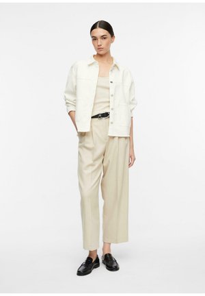 Kobieta stojąca, ubrana w kremową kurtkę, beżowy top na ramiączkach, plisowane beżowe spodnie z czarnym paskiem i czarne mokasyny na białym tle.