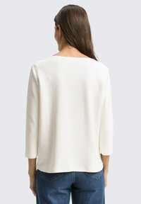 Weißes, strukturiertes Langarmshirt mit quadratischem Ausschnitt, von hinten betrachtet. Kombiniert mit blauen Jeans.