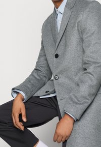 Uomo seduto con un coat di lana grigia sopra una camicia celeste e pantaloni neri, mani appoggiate sulle gambe su uno sfondo semplice.