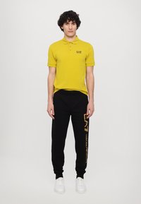 Sárga polo ing három gombbal és EA7 logóval. Fekete melegítőnadrág narancssárga márkázással és szegélyekkel. Fehér sportcipők teszik teljessé az öltözetet.