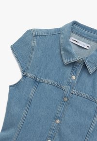 Camisa de denim azul clara de manga corta, con cierre de botones y cuello. Presenta costuras en contraste y una etiqueta que dice "Katan Jeans 40."