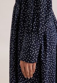 Abito blu navy con pois bianchi, caratterizzato da un scollo arricciato e maniche lunghe a sbuffo. Maniche con polsini e dettaglio a bottone. Tessuto morbido.