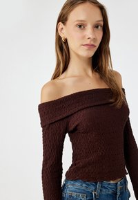 Off-the-shoulder bruine top van textuurstof met een golvend patroon, met lange mouwen en een getailleerde uitstraling. Gecombineerd met blauwe denimshorts.