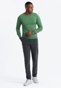 Grüner, gerippter Pullover mit langen Ärmeln, graue maßgeschneiderte Hose und weiße Sneakers. Uhr-Zubehör am Handgelenk sichtbar. Einfache, moderne Freizeitlook.