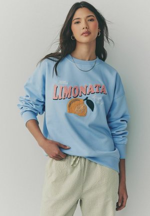 Sweatshirt bleu deschis cu textul "Limonata" și grafică cu lămâie; are mâneci lungi și manșete ribbate, purtat peste pantaloni ușor carotați.