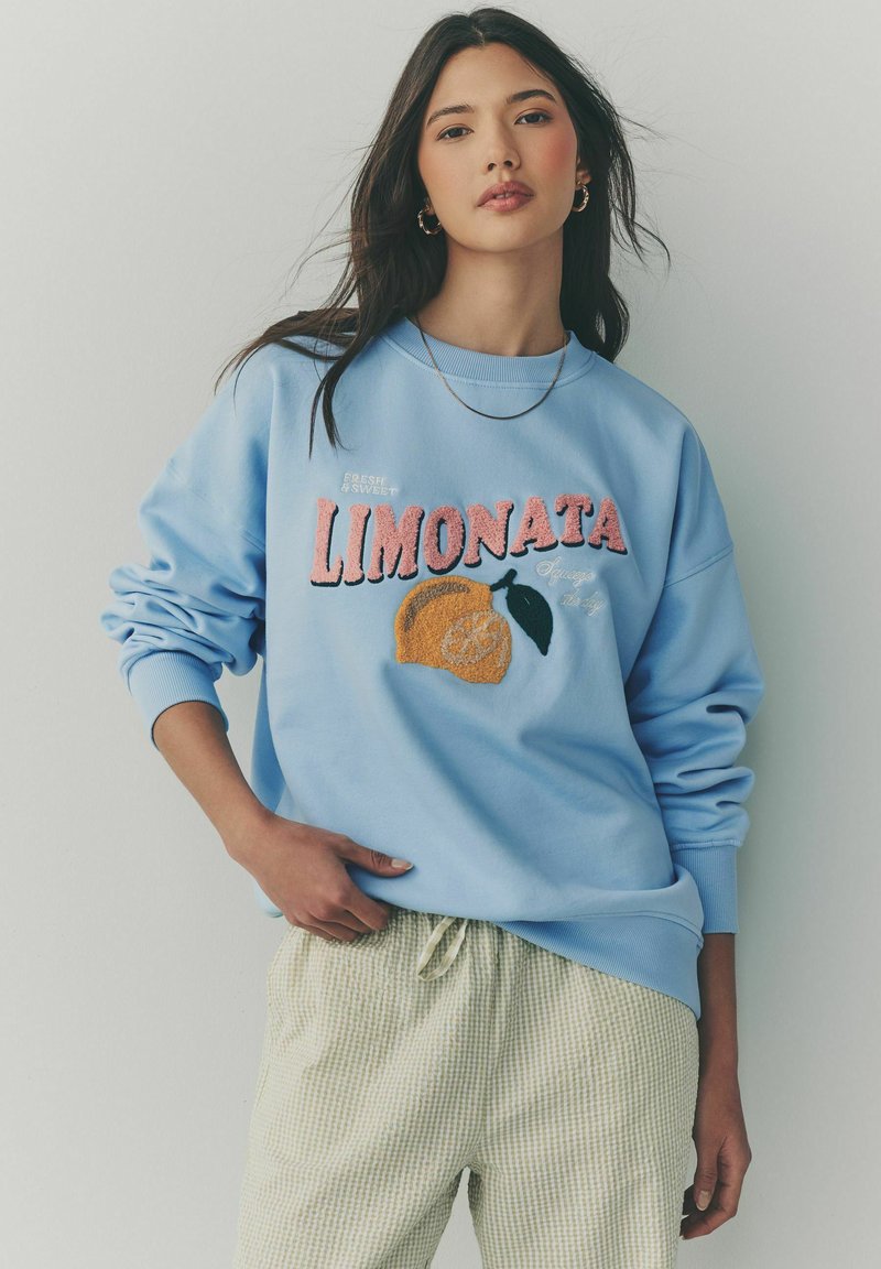 Hellblaues Sweatshirt mit dem Text "Limonata" und einer Zitronengrafik; hat lange Ärmel und gerippte Bündchen, getragen über hellgemusterten Hosen.