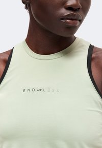 Gros plan d'une personne portant un haut sans manches vert clair avec le mot "ENDLESS" et un symbole d'infini sur la poitrine.
