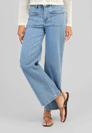 Femme portant un jean bleu clair à jambes larges avec une bordure tressée sur les poches avant et des sandales marron à brides, sur fond gris uni.