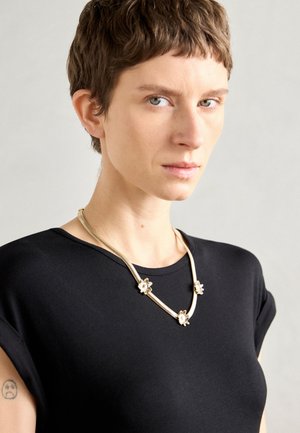 Collier floral en or avec trois accents floraux, chaîne texturée, porté sur une chemise noire à manches courtes sur fond neutre.