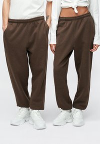 Καφέ φόρμα σε άνετη εφαρμογή με ελαστική μέση και πλαϊνές τσέπες, σε συνδυασμό με άσπρα αθλητικά παπούτσια. Μαλακή υφή, αθλητικός σχεδιασμός.