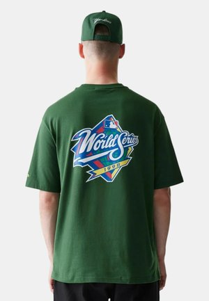 Uomo con cappellino verde e maglietta verde scuro con logo della Serie Mondiale MLB del 1998 sul retro, in piedi davanti a uno sfondo bianco.