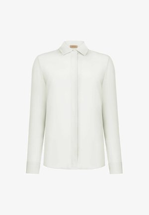 Blusa bianca a maniche lunghe con chiusura frontale con bottoni nascosti, collo a punta e orlo dritto, esposta su sfondo bianco.