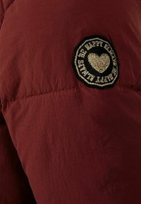 Bordo puffer jakna s glatkom teksturom, s kružnom crnom tkaninom zakrpom sa srcem i tekstom izvezenim zlatnom bojom.