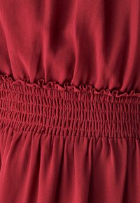 Tkanina crvene haljine ima meku, teksturiranu površinu s pojasom od rastezljive elastike naboranom i volanima na vrhu struka.