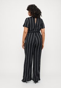 Black jumpsuit with white vertical stripes, short sleeves, back keyhole detail, wide legs, and a tied belt at the waist. Smooth fabric texture.