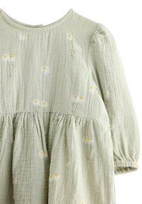 Lichtgroene jurk gemaakt van een textuurstof, met een gerimpelde taille en bloemborduursels met witte madeliefjes en gele accenten.