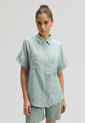 Kobieta z kręconymi włosami upiętymi w kok, ubrana w jasnorozieloną koszulę na krótkie rękawy z guzikami oraz dopasowane szorty, stojąca na jednolitym tle.