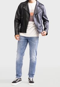 Chaqueta de motorista de cuero negro con cremalleras plateadas sobre una camiseta de rayas clara, combinada con jeans azules deslavados y zapatillas negras.