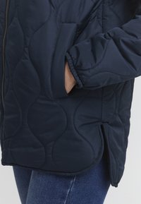 Marineblå quiltet jakke med en glat tekstur, der har lynlåslukning og sidelomme. Manchetterne er elastiske, og kanten er let buet.