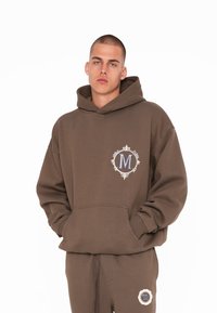 Brun hættetrøje lavet af blødt stof, med et stort broderet logo i cirkulært design på brystet og en lomme foran.