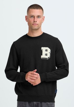 Mand med kort hår iført en sort sweatshirt med et stort hvidt bogstav "B" på brystet, stående med hænderne samlet.