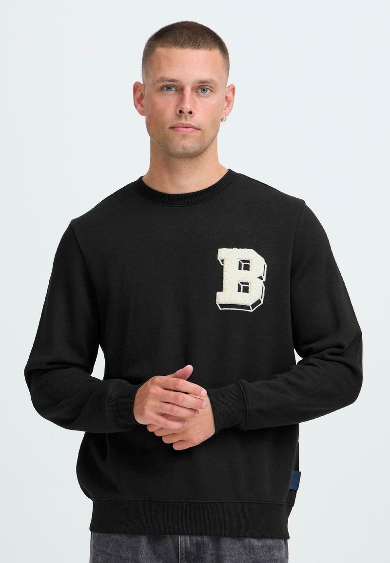 Hombre con cabello corto que lleva una sudadera negra con una gran letra blanca "B" en el pecho, de pie con las manos juntas.