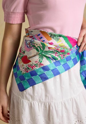 Frau trägt ein pink geripptes Oberteil und einen weißen Rock mit einem bunten Schal, der Häuser, Bäume, Blumen und einen blau karierten Rand zeigt und um die Taille gebunden ist.