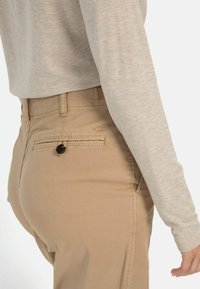 Beige Hose mit slim fit, ausgestattet mit einer Gesäßtasche mit Knopfverschluss, kombiniert mit einem hellgrauen Langarmoberteil. Weiche Textur, lässiges Design.