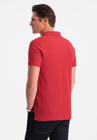 Polo rouge à manches courtes, avec un col classique et une texture lisse. Porté avec un jean foncé, montrant un design simple et épuré.