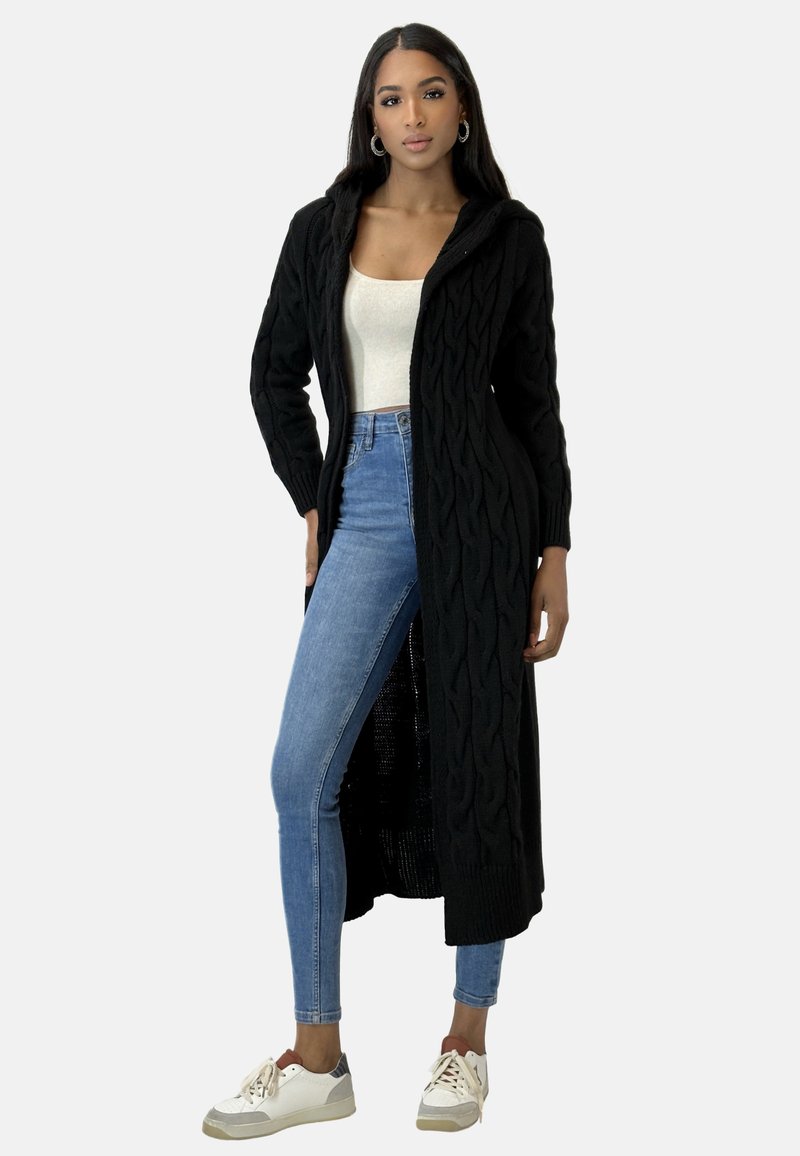 Svart stickad cardigan med huva, med ett kabelmönster. Bärs över en krämfärgad topp och kombineras med blå denim och vita sneakers.