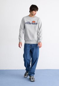 Grijze sweatshirt met "ellesse" logo, ronde hals en geribbelde manchetten, gecombineerd met blauwe denim jeans en sportschoenen met zwarte accenten.