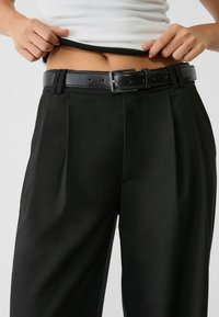 Pantalons noirs ajustés avec taille haute, plis, et une ceinture en cuir brillant. Le tissu est lisse avec une légère brillance et une coupe structurée.
