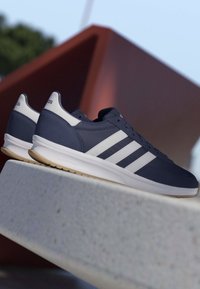 Par de zapatillas Adidas azul marino con rayas blancas y suelas de goma descansando sobre una superficie de hormigón con una escultura roja abstracta al fondo.