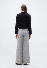 Γυναίκα που στέκεται γυρισμένη προς τα πίσω, φοράει μαύρο cropped σακάκι, γκρι ανοιχτού χρώματος παντελόνι με φαρδιά μπατζάκια και πίσω τσέπη, και σκούρα παπούτσια σε απλό φόντο.