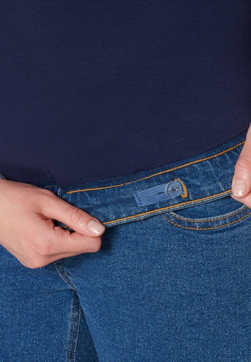 Jean en denim bleu avec des coutures oranges, présentant un bouton de fermeture et une étiquette. La taille est visible avec une main qui la tient.