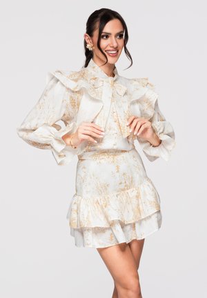 Kvinde smiler, iført en hvid blomsterprintet bluse med flæser og sløjfe samt en matchende flerlags mininederdel, poserer mod en ensartet lys baggrund.