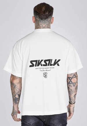 Mężczyzna z tatuażami na szyi i ramieniu, ubrany w białą koszulkę SikSilk z czarnym logo i napisem na plecach, stojący na jednolitym tle.