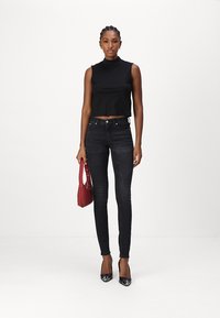 Top negro cortado sin mangas, jeans ajustados oscuros y tacones negros de punta. Sosteniendo un bolso rojo pequeño con una correa y acentos de tachuelas.