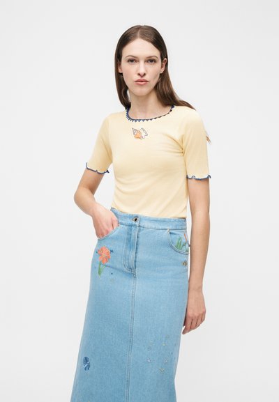 Γυναίκα που φοράει ανοιχτό κίτρινο τοπ με κέντημα με κοχύλια και ανοιχτή μπλε τζιν φούστα με λουλουδάτο και αστερόμορφο κέντημα, στέκεται μπροστά σε λευκό φόντο.