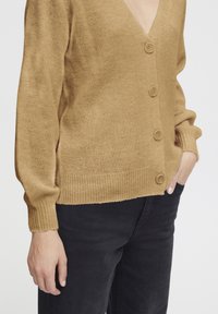 Strickjacke in Camel-Farbe mit tiefem V-Ausschnitt, fünf Knöpfen, gerippten Bündchen und Saum, kombiniert mit schwarzen Hosen.