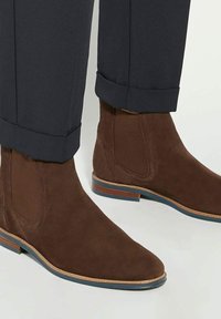 Bottines en daim marron avec un bout pointu, des panneaux latéraux élastiques et une semelle rayée bleu et marron contrastante, associées à un pantalon sombre.
