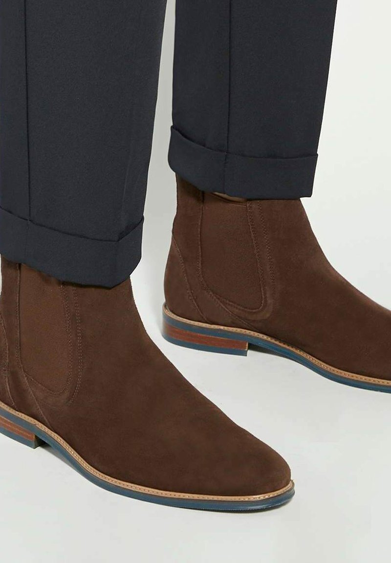 Bottines en daim marron avec un bout pointu, des panneaux latéraux élastiques et une semelle rayée bleu et marron contrastante, associées à un pantalon sombre.