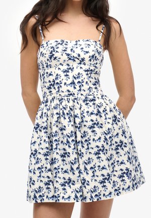 Kobieta w białej sukience bez rękawów z cienkimi ramiączkami i niebieskim kwiatowym wzorem, stojąca z rękami za plecami.