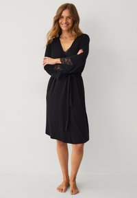 Robe noire en tissu doux, avec des accents en dentelle sur les manches évasées et une taille cintrée avec une ceinture, de conception mi-genoux.